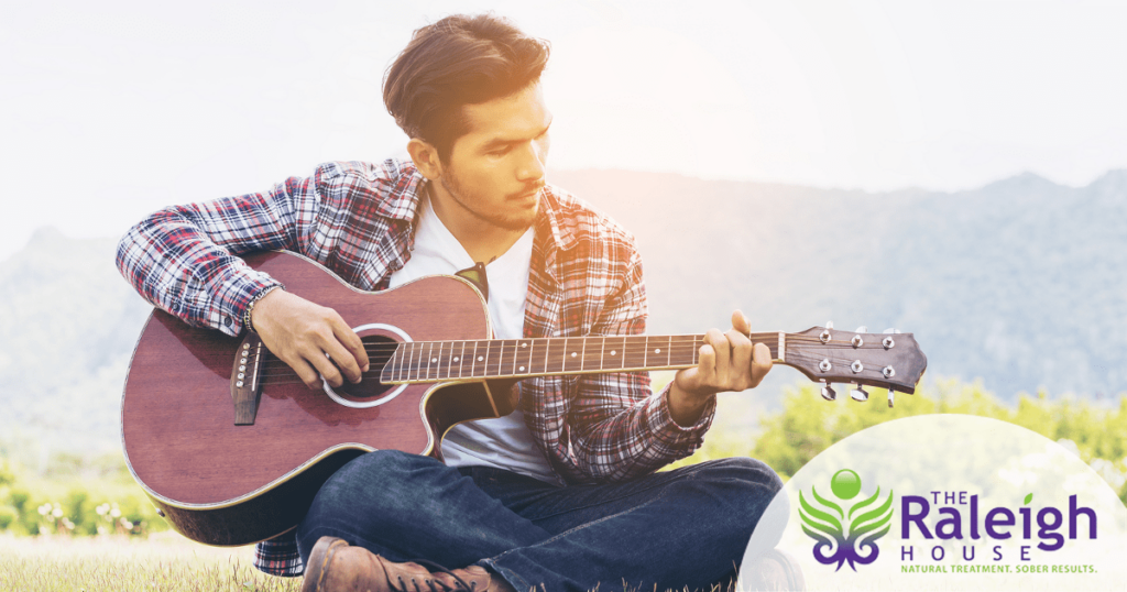 An attractive young man sits on the grass, playing his guitar.