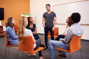 A man standing up and addressing a group of people in a therapy session.