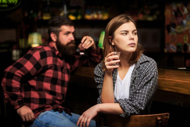 A caucasian woman drinking at a bar with a caucasian male watching her in the background.
