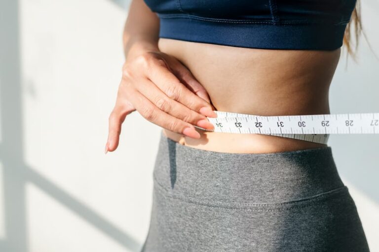Woman in sports bra and workout pants measuring her waist with measuring tape.