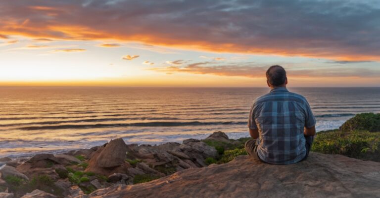 man sits by the ocean and watches the sunset.